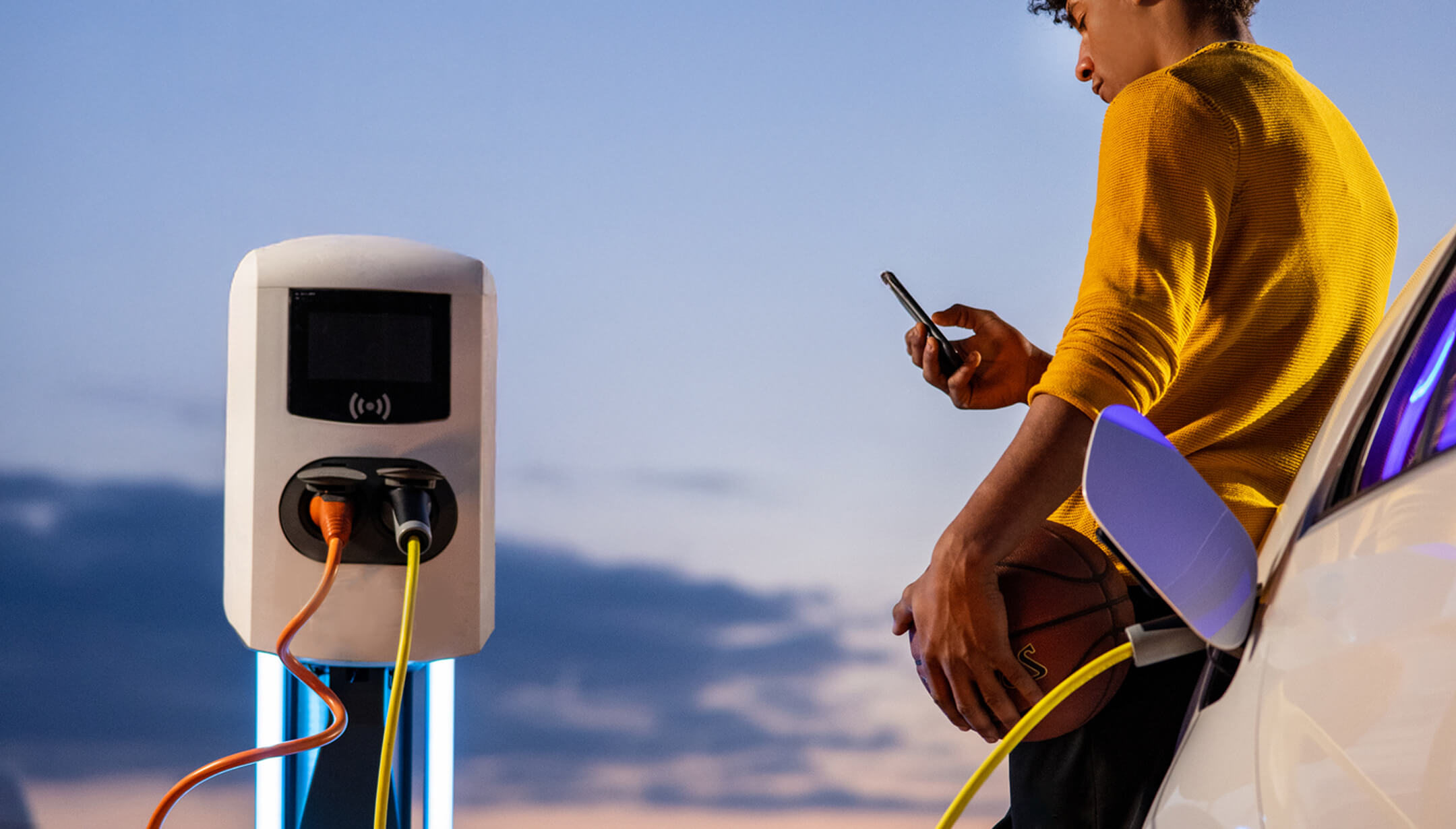 Un garçon avec un portable et un ballon de basket entre une borne électrique et une voiture en charge